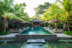Une piscine entourée de palmiers, avec des chaises longues et des parasols, et un petit bâtiment au centre.
