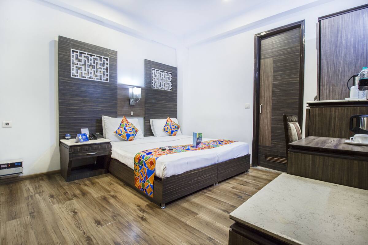 A hotel room with a wooden floor, a bed with a colorful blanket, a nightstand, a desk, and a wardrobe.