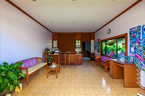 A reception area with a wooden reception desk, a sofa, a small table with a vase, and a large window with a view of greenery. - My Village Lamai (Koh Samui)