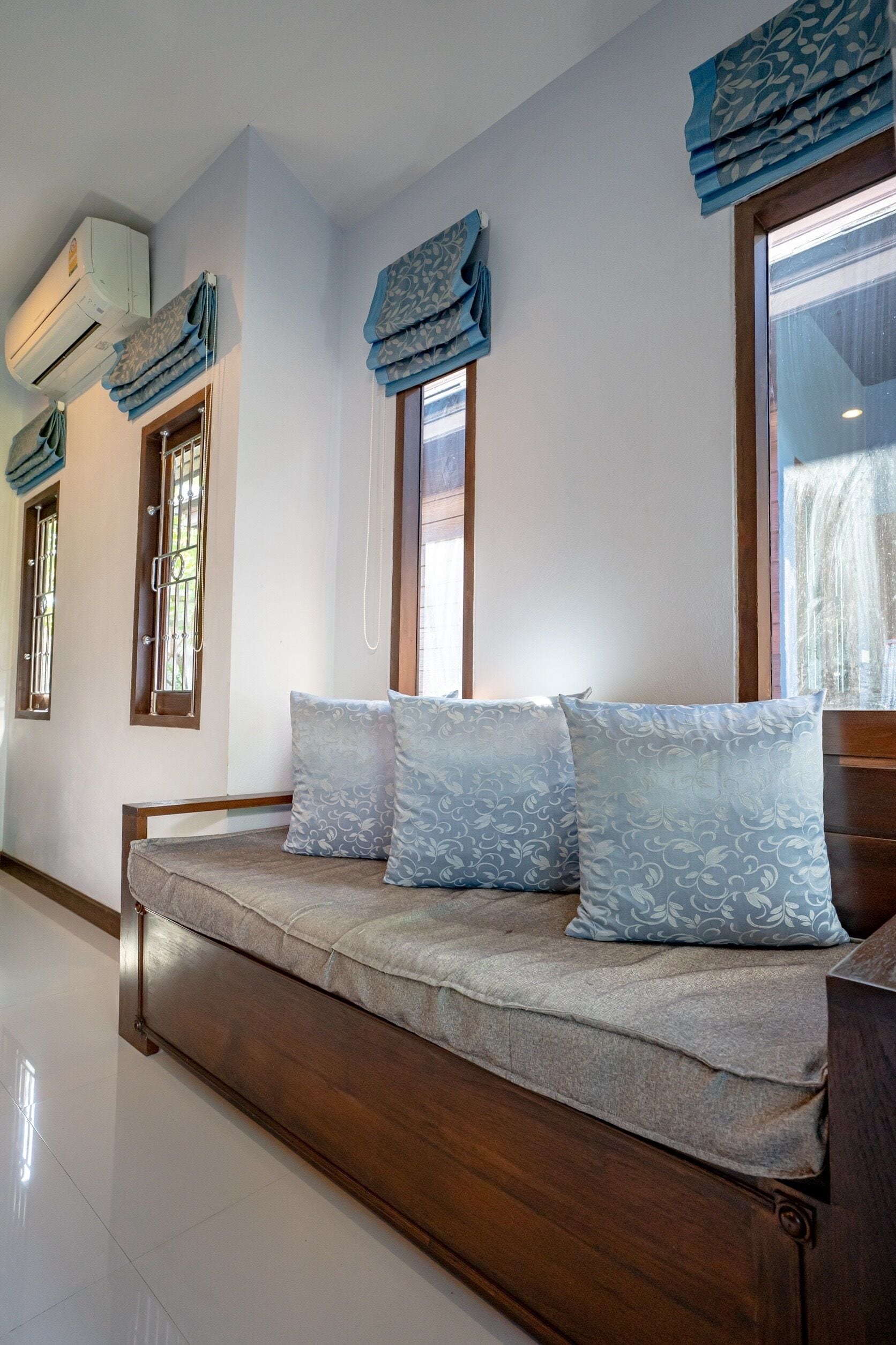 Quarto com um banco de madeira com almofadas, uma janela com cortina azul e um ar condicionado fixado na parede.