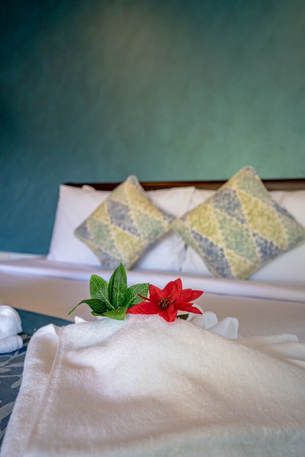 Una cama bien hecha con sábanas blancas, dos almohadas decorativas con un diseño geométrico y una flor roja con hojas verdes sobre la colcha.