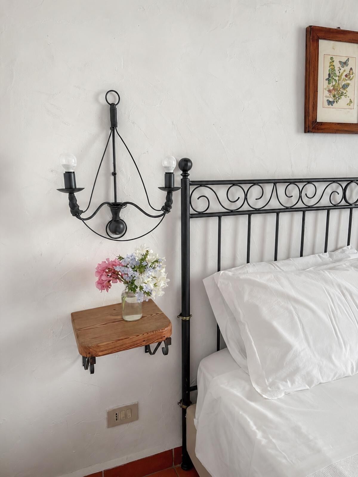 Un letto con biancheria bianca, una testiera in metallo nero, una mensola a parete con un vaso di fiori e un applique nero con due lampadine.