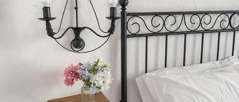 A bed with white bedding, a black metal headboard, a wall-mounted shelf with a vase of flowers, and a black wall sconce with two bulbs.
