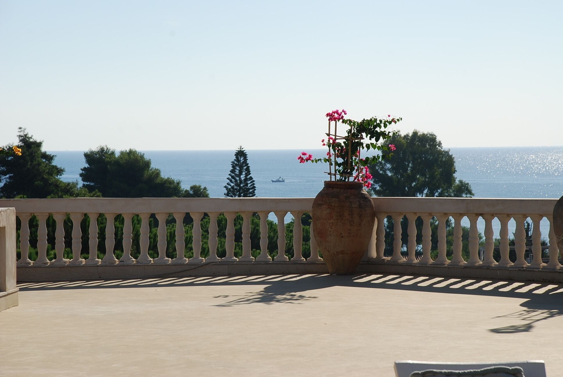 Ein Balkon mit einer großen Vase voller pinkfarbener Blumen, einem Geländer und Blick aufs Meer.
