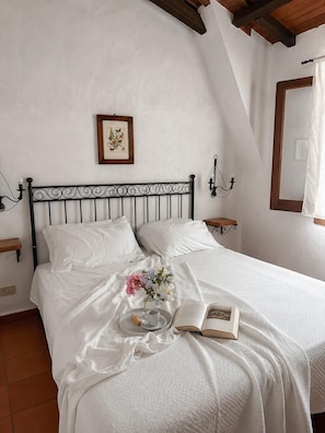 A bedroom with a bed, a bedside table, a wall-mounted light, a framed picture, and a window with curtains.