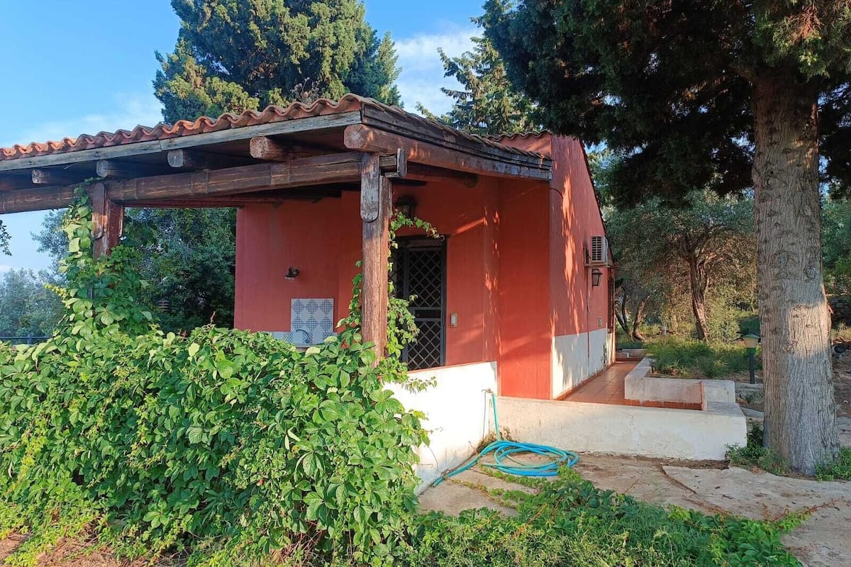 A small, red building with a tiled roof, surrounded by greenery and trees.