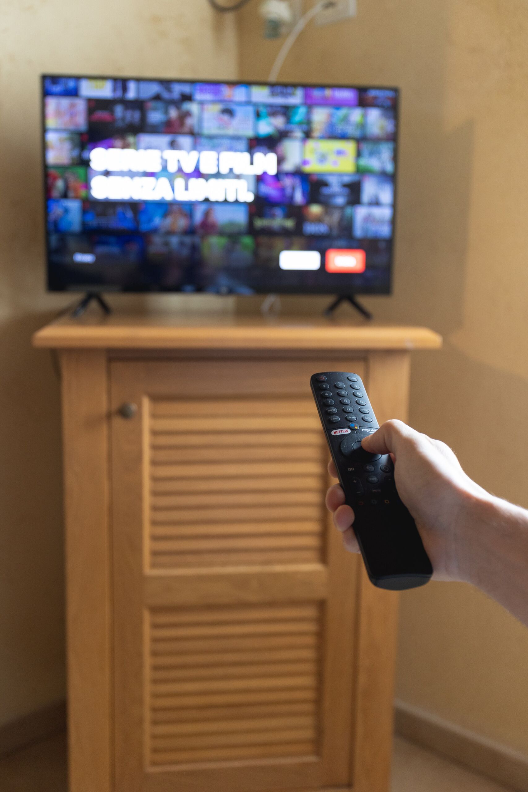 A hand holding a remote control in front of a television displaying multiple channels.