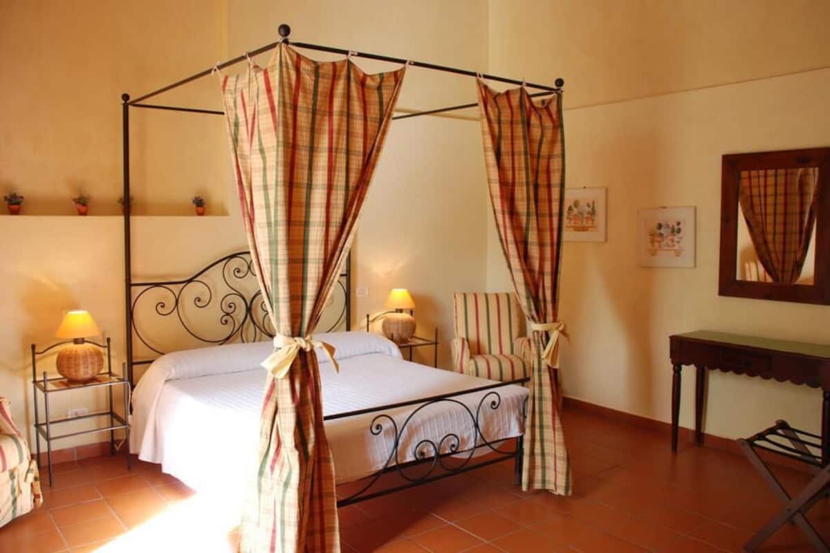 A four-poster bed with striped curtains, a bedside table with a lamp, a chair, and a small table with a mirror.