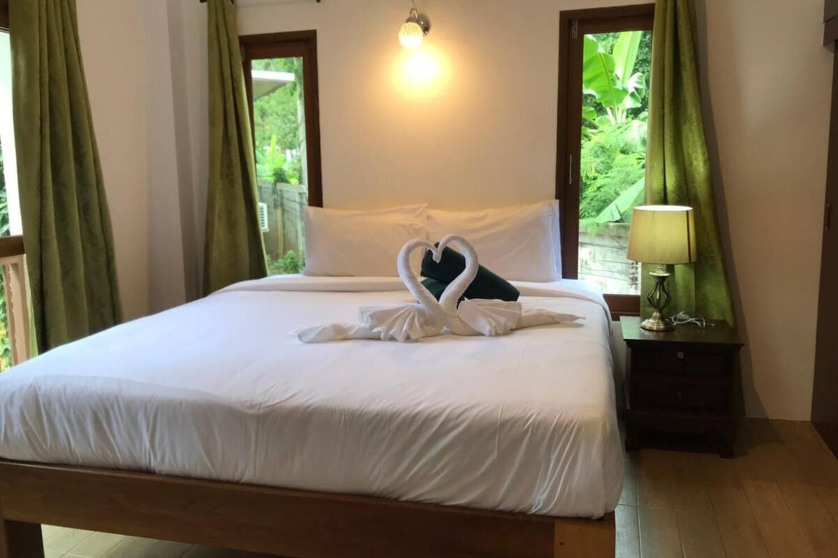 Un lit parfaitement fait, avec du linge de lit blanc et une serviette pliée en forme de cygne. Une lampe de chevet est posée sur une table de nuit en bois.
