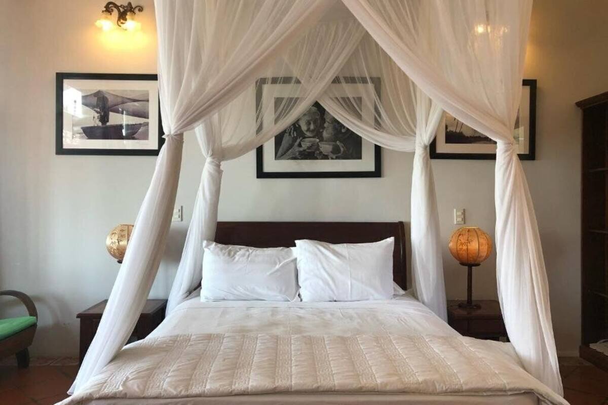 Una cama con ropa blanca y dosel, cuadros en la pared y una lámpara en la mesita de noche.