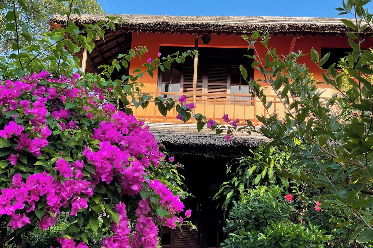 Una casa tradicional con techo de paja, balcón y un jardín lleno de coloridas flores de buganvilla.