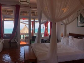 A bedroom with a bed, a nightstand, a chair, and a view of the ocean through large windows.