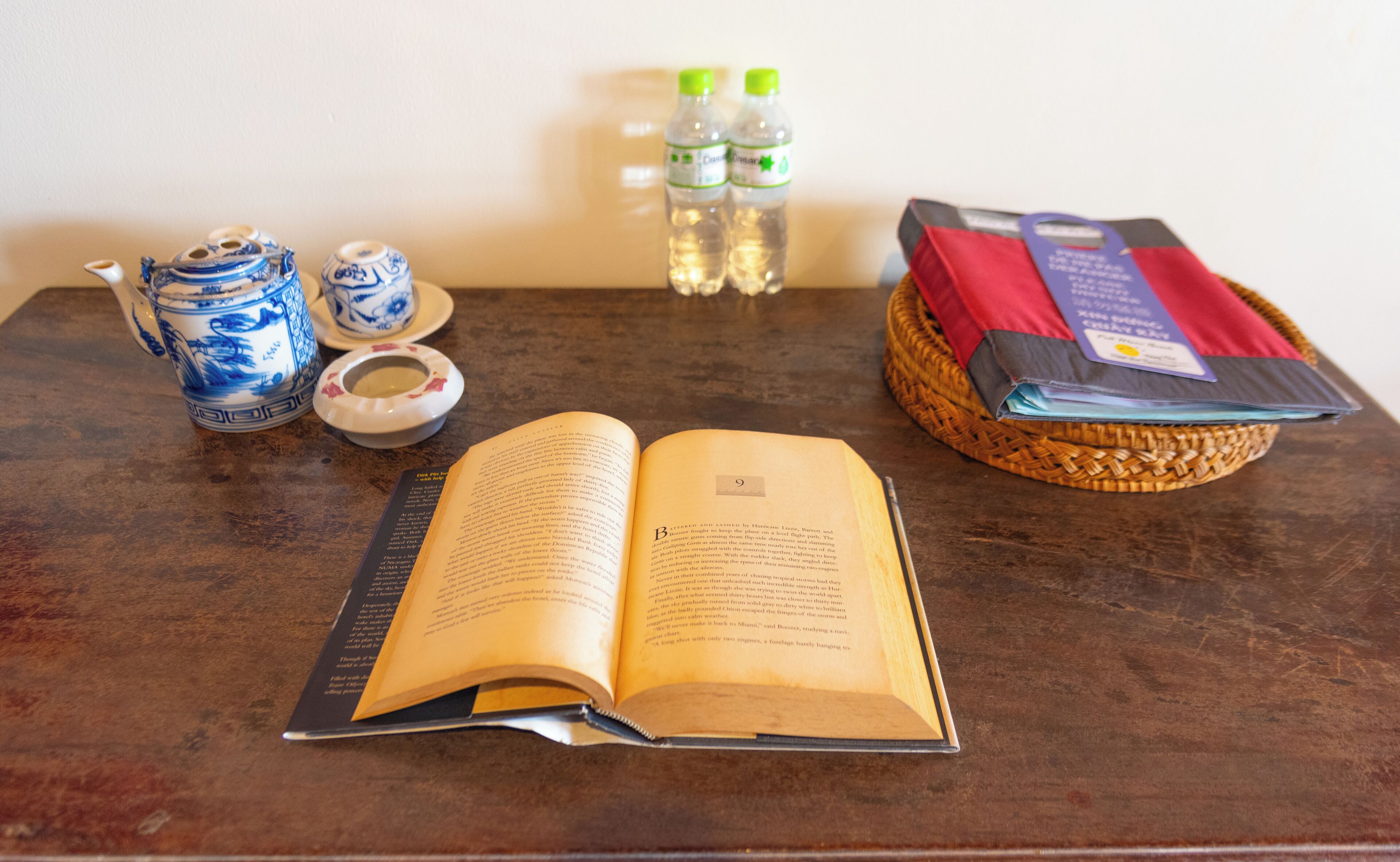 A book open on a table, a teapot, a cup, two bottles of water, and a basket with a red item.