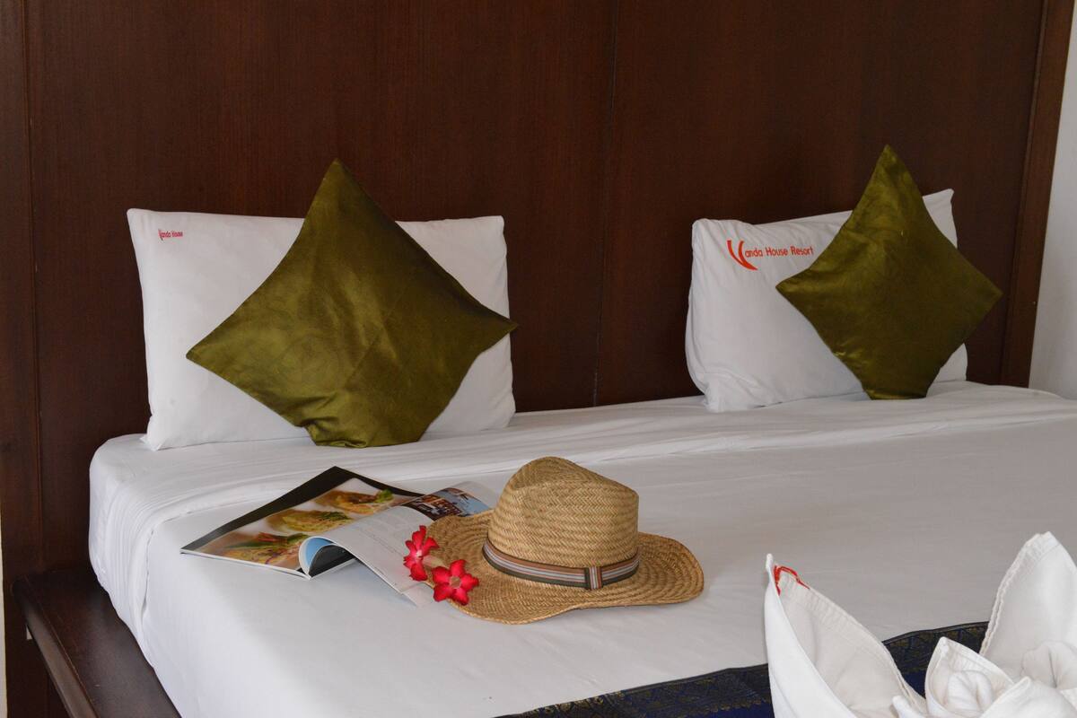 A neatly made bed with white linens and green pillows, a straw hat, and a magazine on the bed.