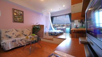 A modern living room with a sofa, a glass coffee table, a TV, and a large window offering a city view.