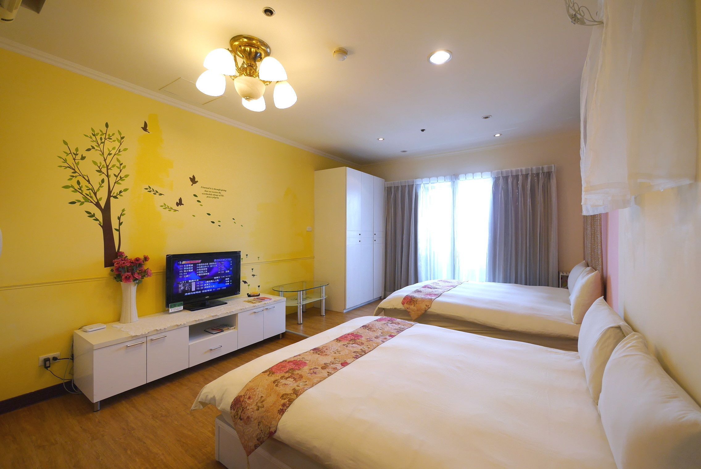 Quarto de hotel com duas camas, uma televisão em um console branco, um vaso com flores e um mural na parede.
