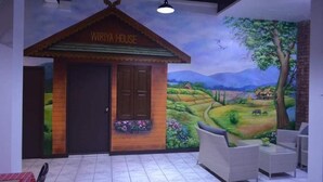 A cozy room with a mural of a countryside scene, a small wooden hut labeled "WIRYA HOUSE," and comfortable seating. - Wiriya House (Chiang Mai)