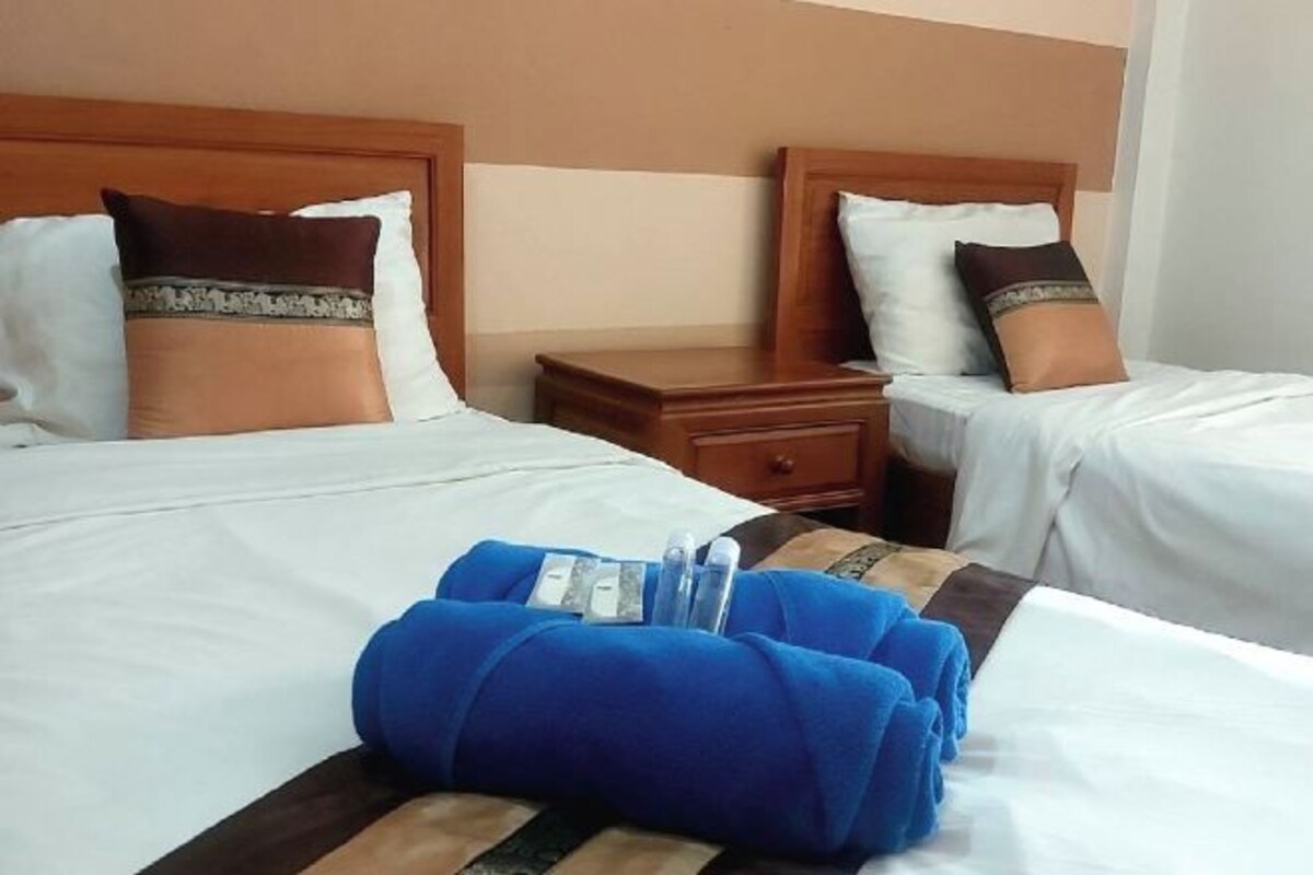 A hotel room with two beds, each with white bedding and brown decorative pillows. There is a wooden nightstand between the beds with a drawer. A blue towel and toiletries are placed on the bed in the foreground.