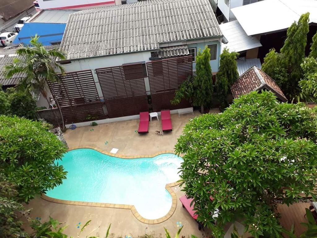 Una piscina con cubierta de madera y sillas de descanso rojas, rodeada de vegetación y un edificio con techo de tejas.