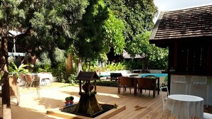 An outdoor patio area with a wooden deck, a small fountain, outdoor seating, and a pool in the background. - Wiriya House (Chiang Mai)