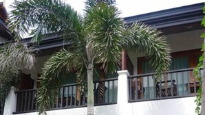 Um edifício de dois andares com uma varanda no andar superior, uma escadaria que leva à entrada e uma pequena área de jardim com palmeiras.