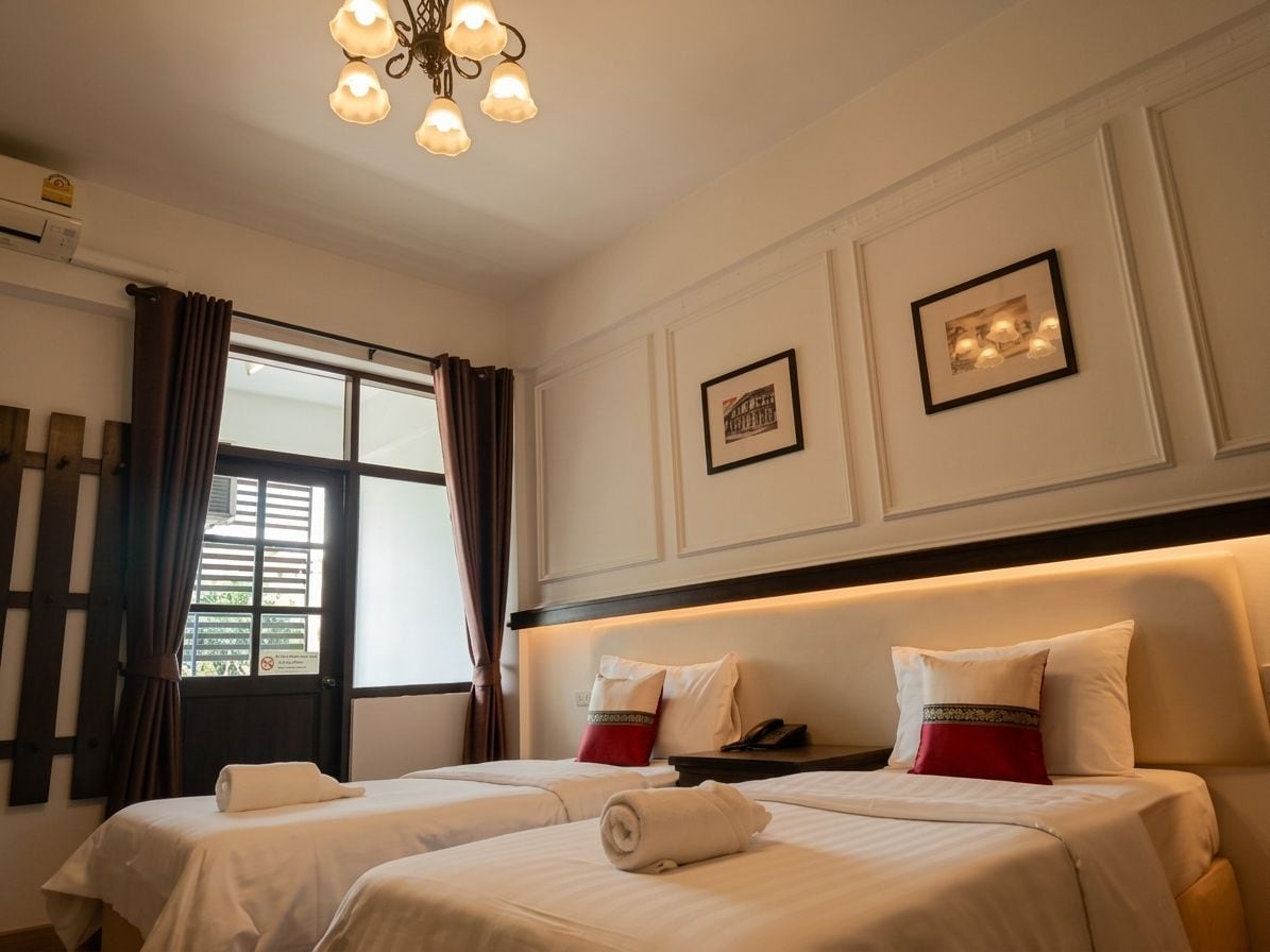 A hotel room with two beds, a large window with curtains, a chandelier, and framed pictures on the wall.