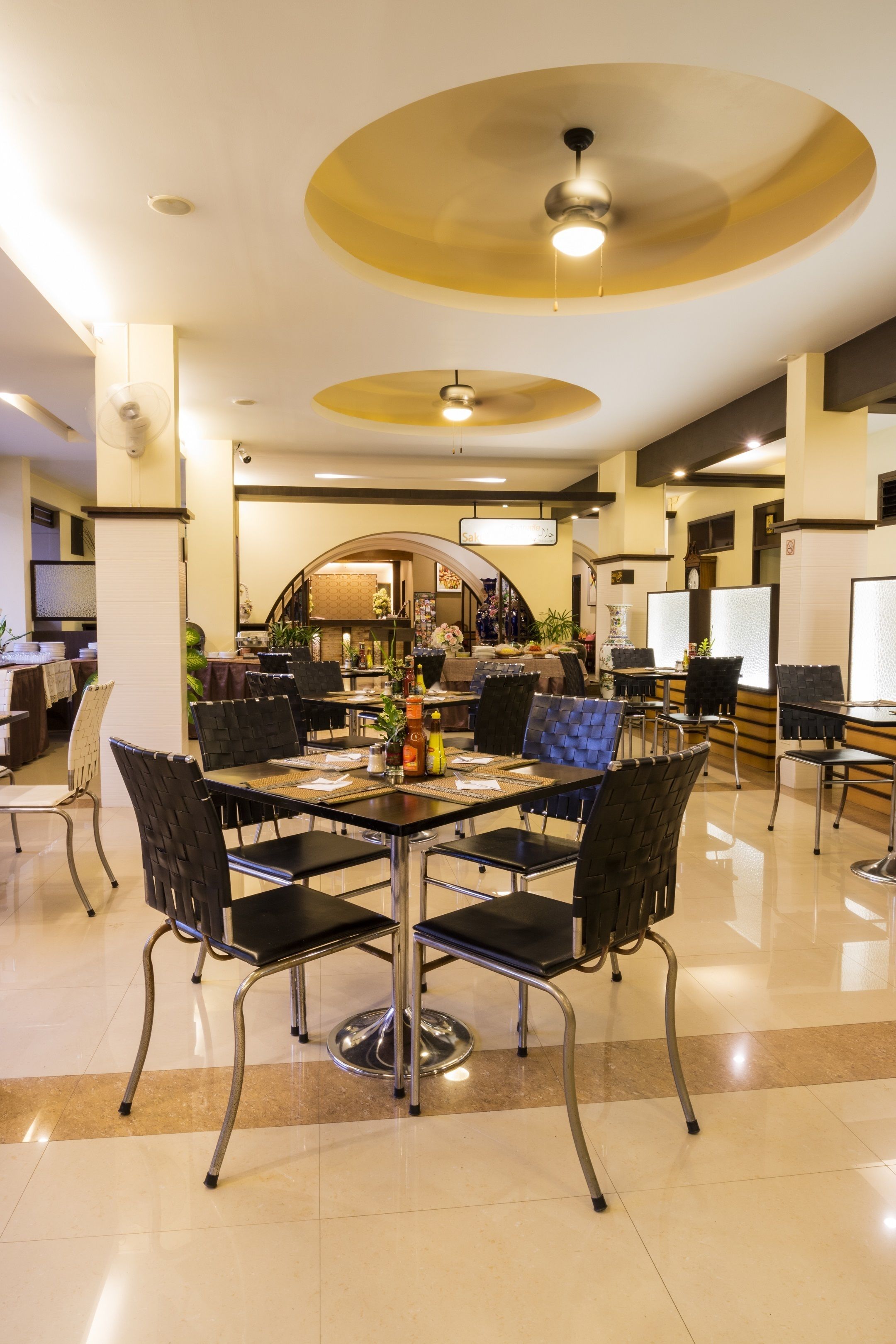 A dining area with a central table surrounded by chairs, a ceiling fan, and a kitchen area in the background.