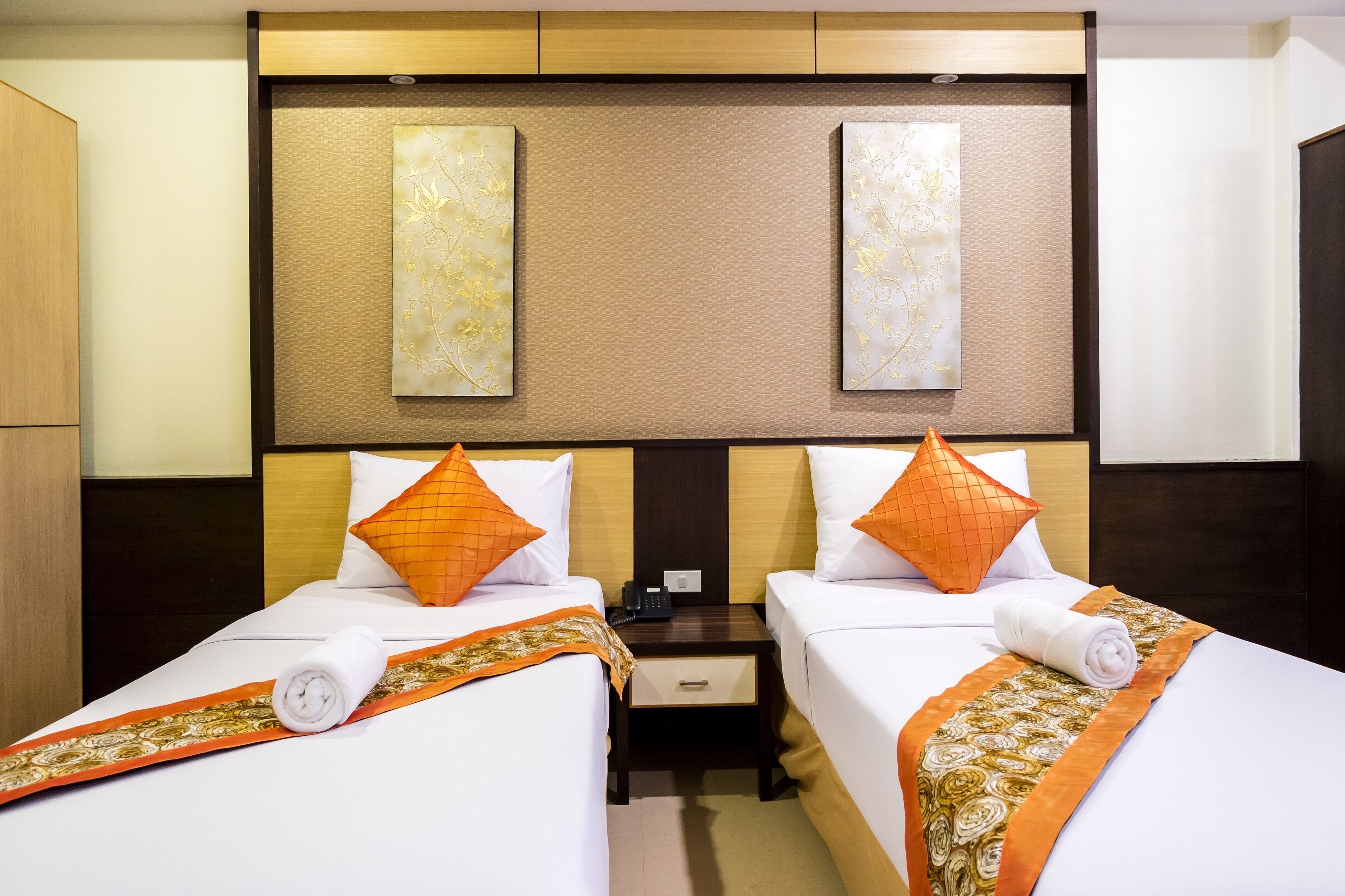 A hotel room with two beds, each with an orange pillow, a wooden headboard, and a small nightstand with a phone.