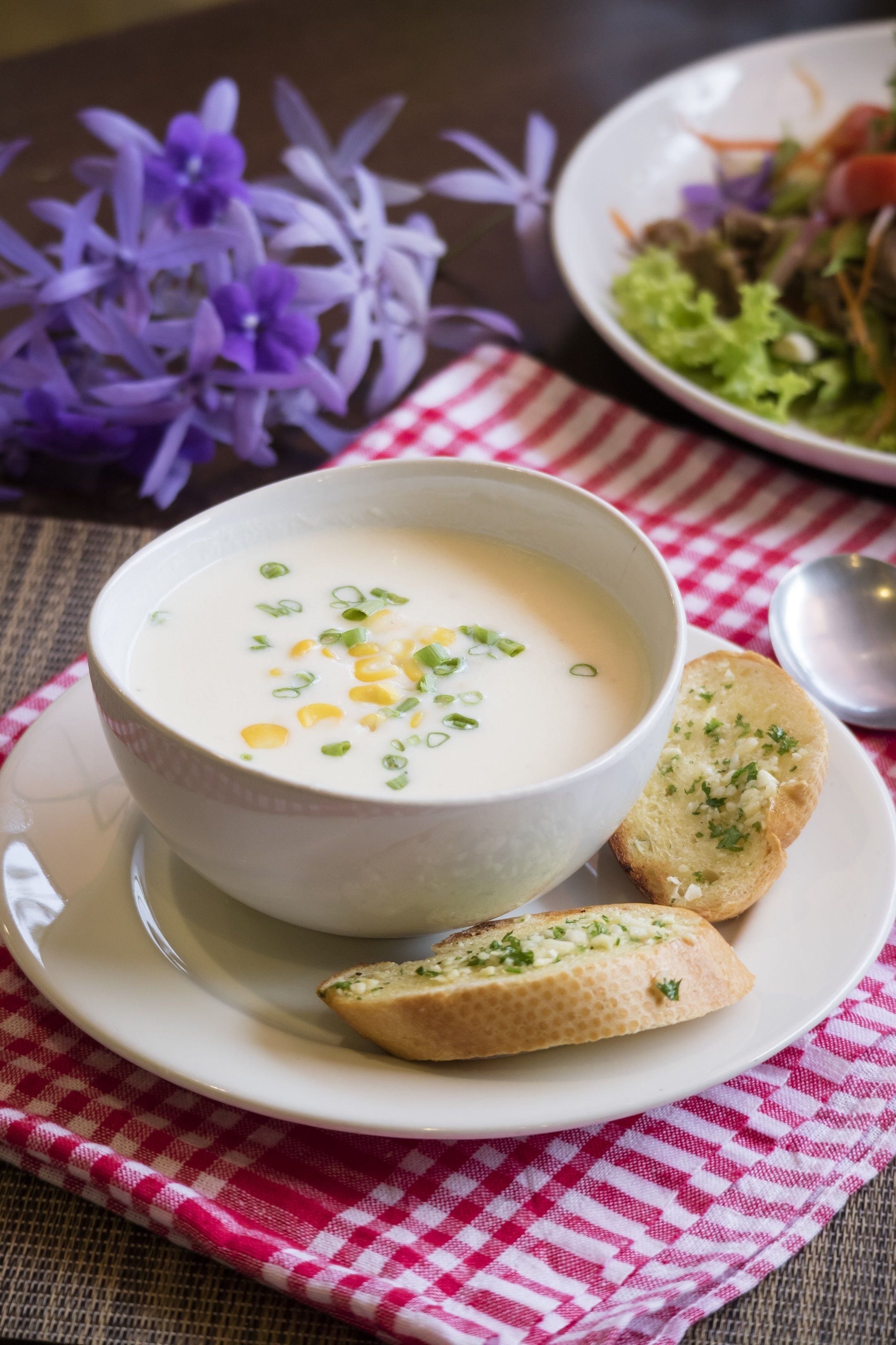 Un bol de soupe aux maïs et aux oignons verts, servi sur une assiette blanche, accompagné de deux morceaux de pain à l’ail, le tout posé sur une serviette à carreaux rouges et blancs.