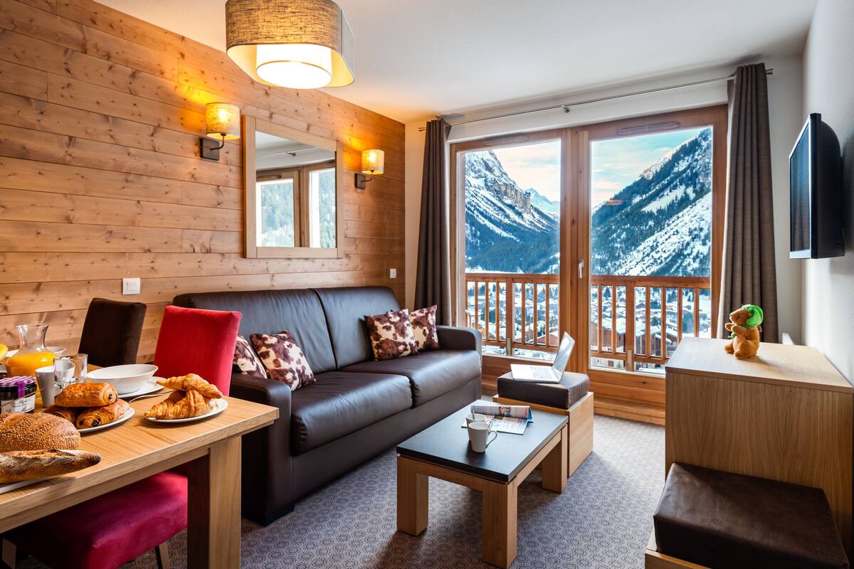 Ein gemütliches Zimmer mit Holzwand, Ledersofa, Esstisch mit Frühstück und einem großen Fenster mit Bergblick.