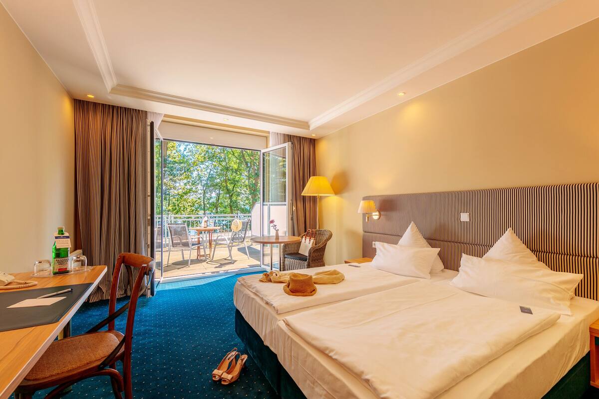 A hotel room with two beds, a desk, a chair, a table, and a view of the outdoors.