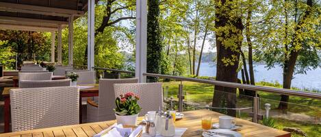 An outdoor dining area with wicker furniture, a wooden table, and a view of a lake surrounded by trees.