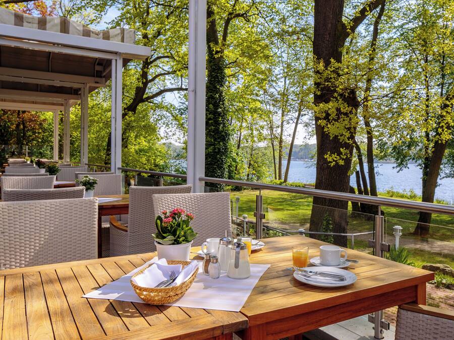Un área de comedor al aire libre con mobiliario de mimbre, una mesa de madera y vistas a un lago rodeado de árboles.