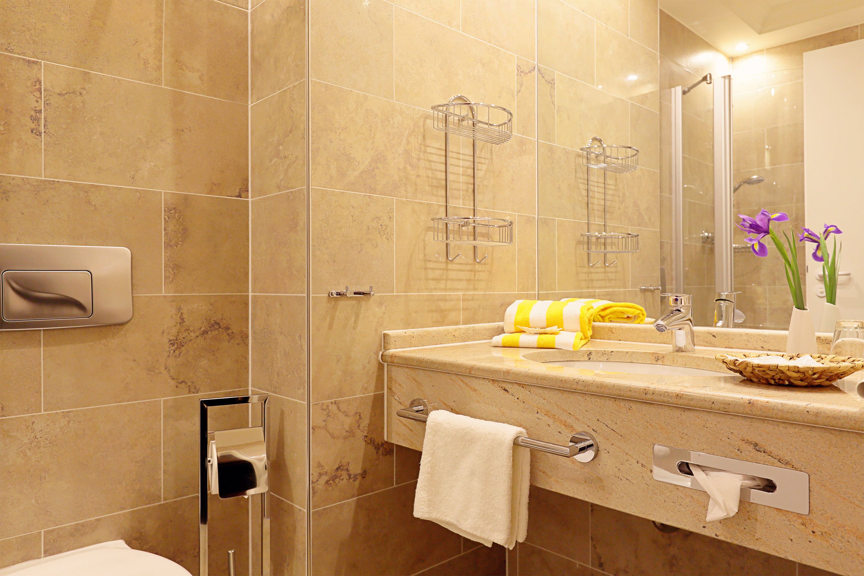 Baño con pared de azulejos de mármol, cabina de ducha de vidrio, lavabo con toallero y espejo con un jarrón de flores.