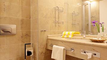 Baño con pared de azulejos de mármol, cabina de ducha de vidrio, lavabo con toallero y espejo con un jarrón de flores.