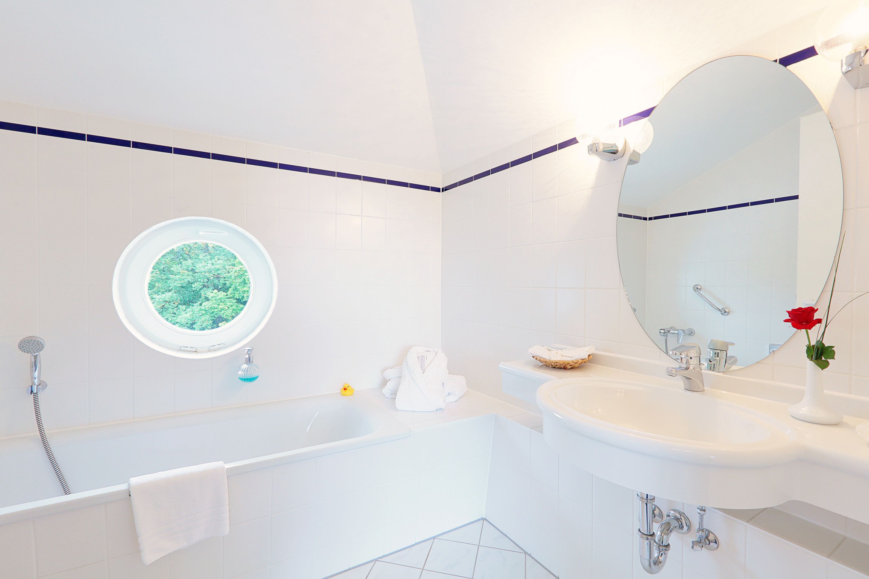 Un baño moderno con bañera, lavamanos, un espejo redondo y ventana con vistas al verdor.