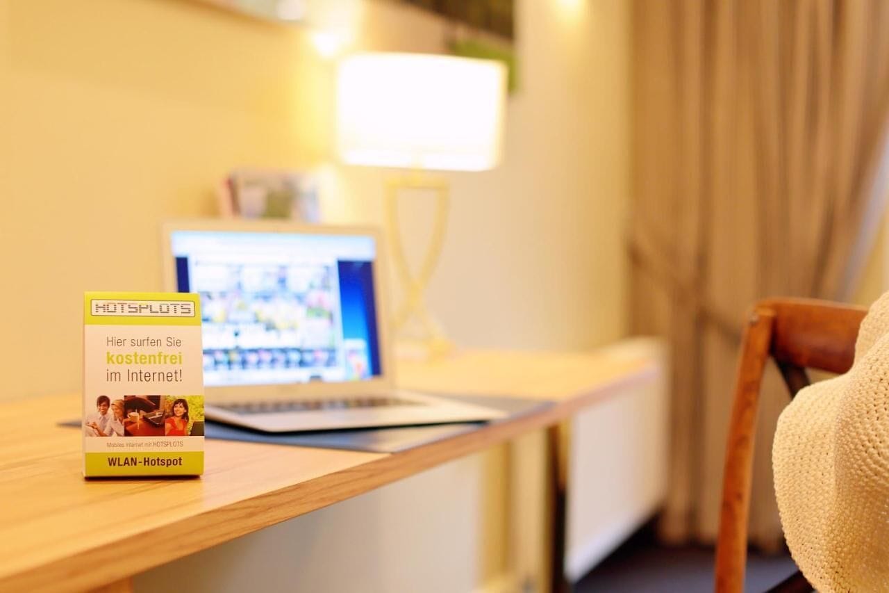 Hotellihuone, jossa on puinen työpöytä, kannettava tietokone, lamppu ja kyltti, jossa kerrotaan WLAN-hotspotin sijainti.