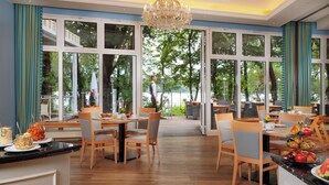 A dining area with wooden tables and chairs, large windows with a view of trees, and a chandelier. - INSELHOTEL Potsdam - Hermannswerder (Potsdam)