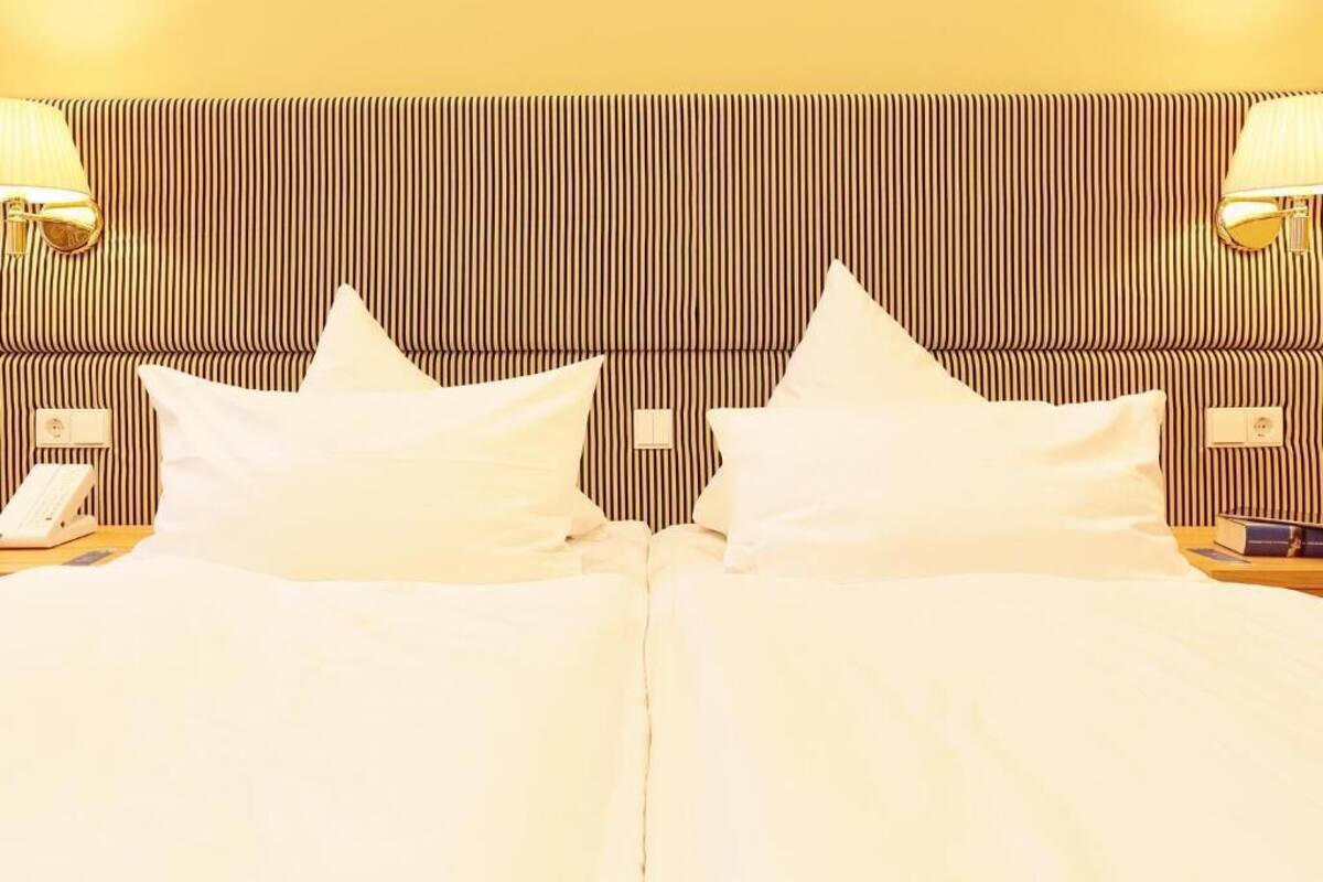A hotel room with a striped headboard, two white pillows, a bedside lamp, a phone, and a book on the nightstand.