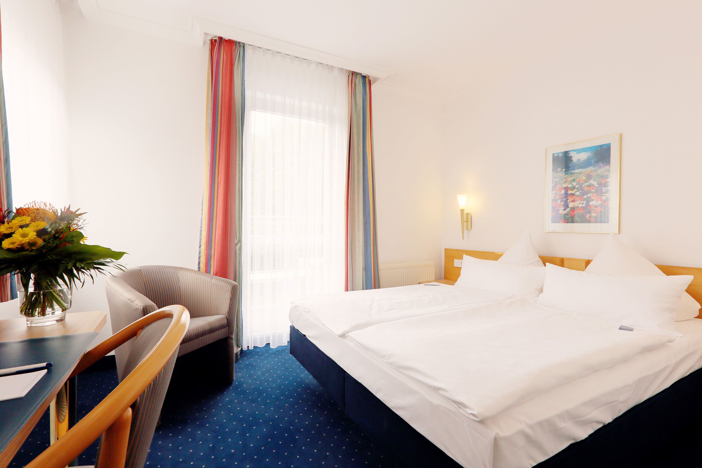 Una habitación de hotel con una cama, un escritorio con silla, un jarrón de flores y un cuadro en la pared.