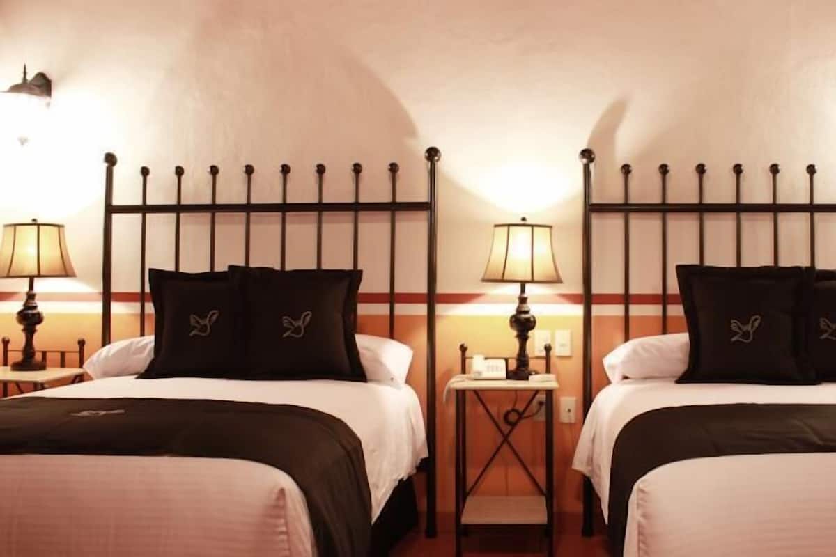 Une chambre d’hôtel avec deux lits, chacun doté d’une tête de lit noire et d’oreillers assortis, une petite table de chevet avec une lampe et un luminaire fixé au mur.