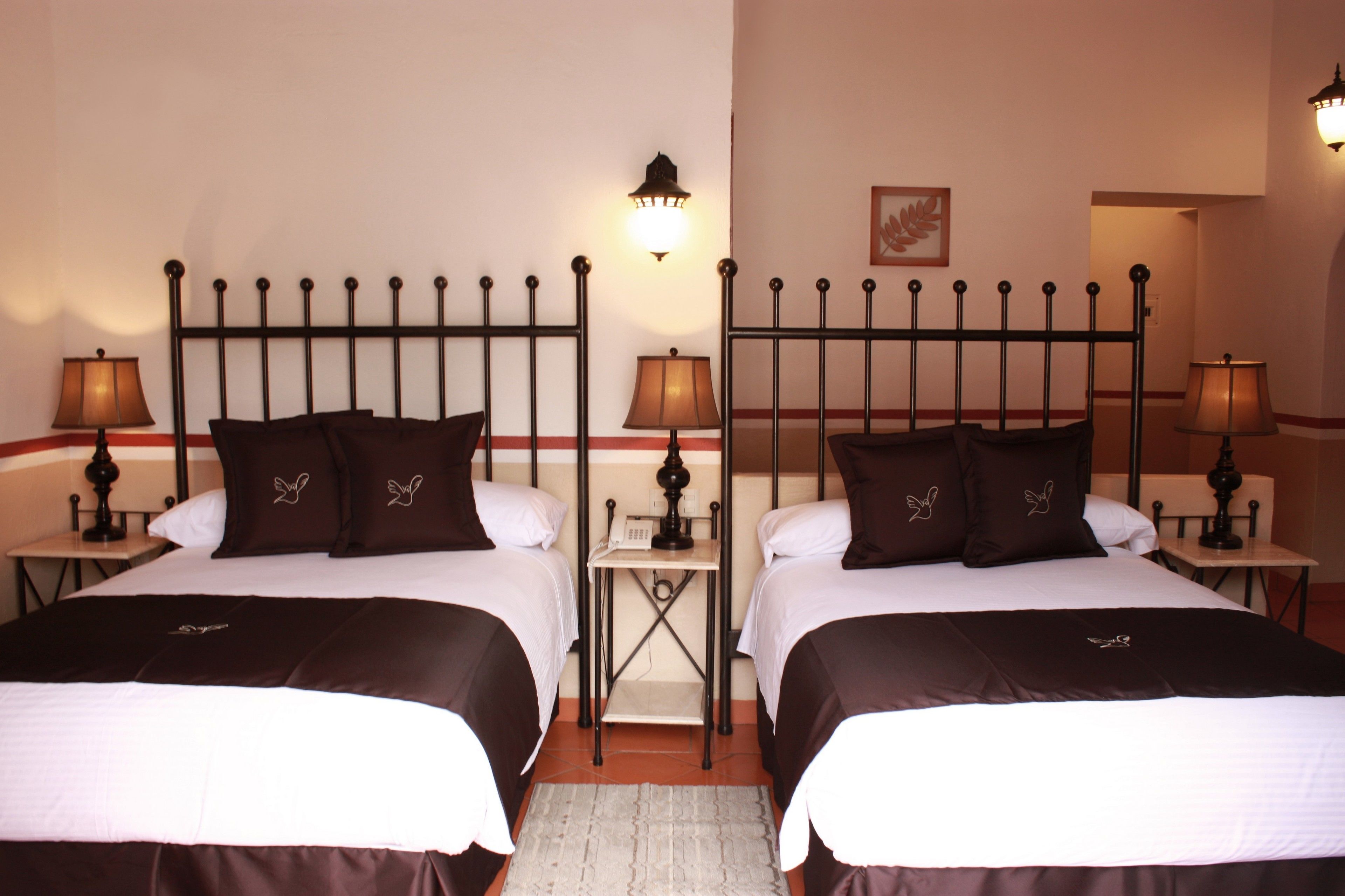 Une chambre d’hôtel avec deux lits, chacun doté d’une tête de lit en métal noir, de draps blancs et d’oreillers marron. On y trouve des tables de chevet avec des lampes, une petite étagère avec un téléphone et un tableau encadré au mur.