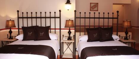 A hotel room with two beds, each with a black metal headboard, white bedding, and brown pillows. There are bedside tables with lamps, a small shelf with a telephone, and a framed picture on the wall.