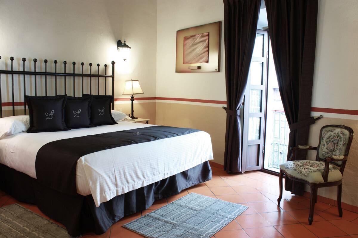 Une chambre d’hôtel avec un lit, une chaise, une lampe, un tapis et une fenêtre avec des rideaux.