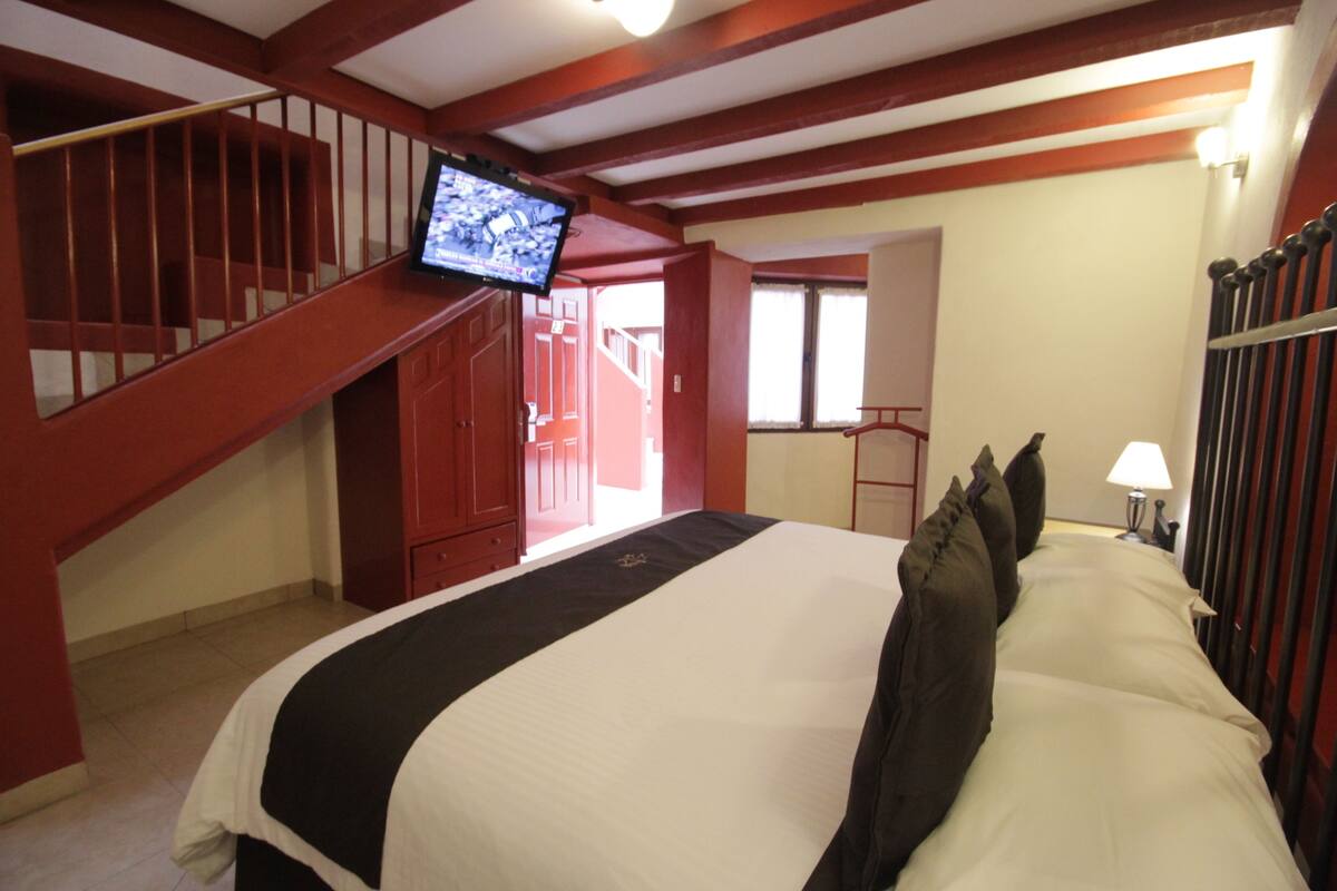 A hotel room with a large bed, a television mounted on the wall, a staircase, and a window.