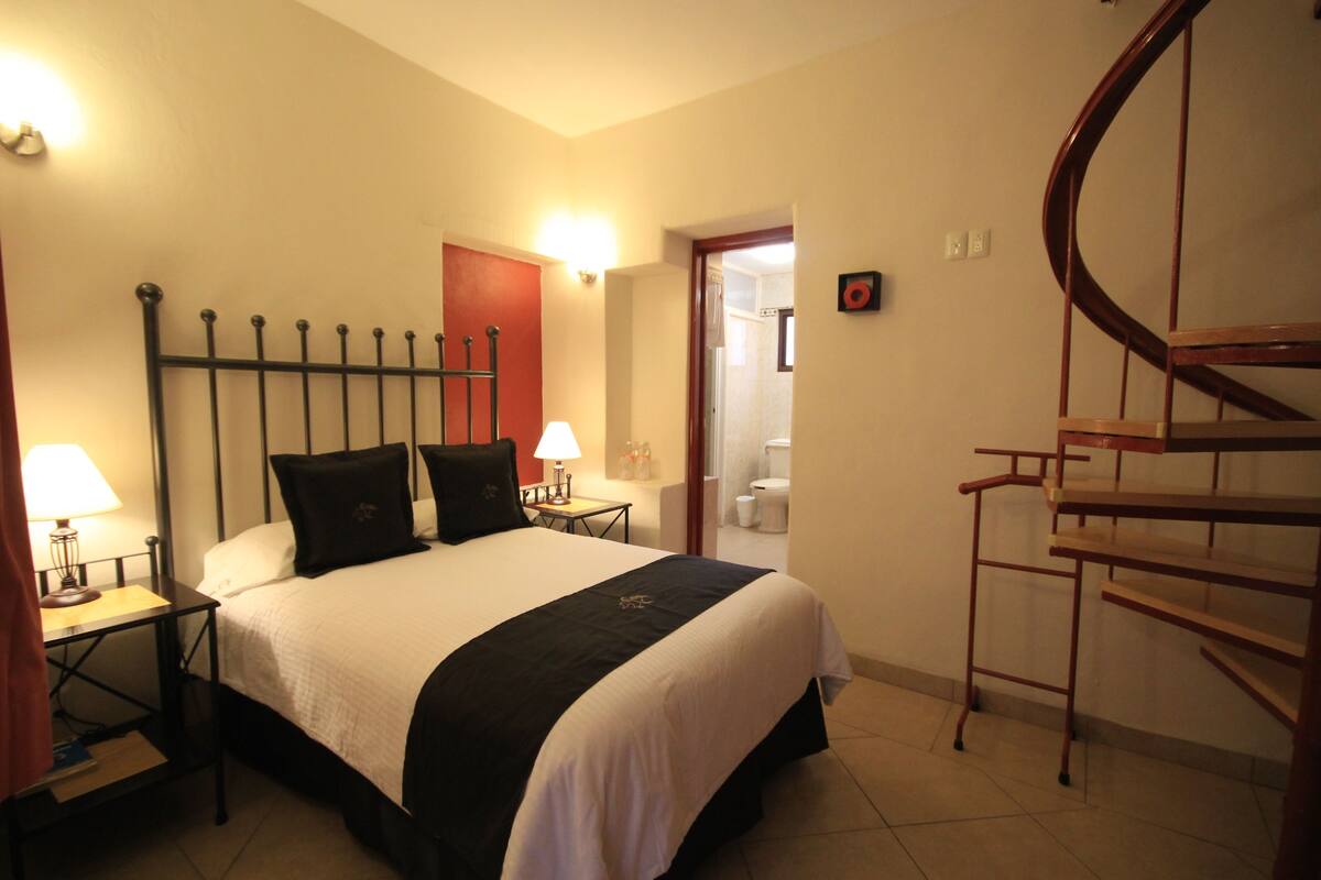 A hotel room with a bed, nightstands, a lamp, a red door, a bathroom, and a wooden spiral staircase.