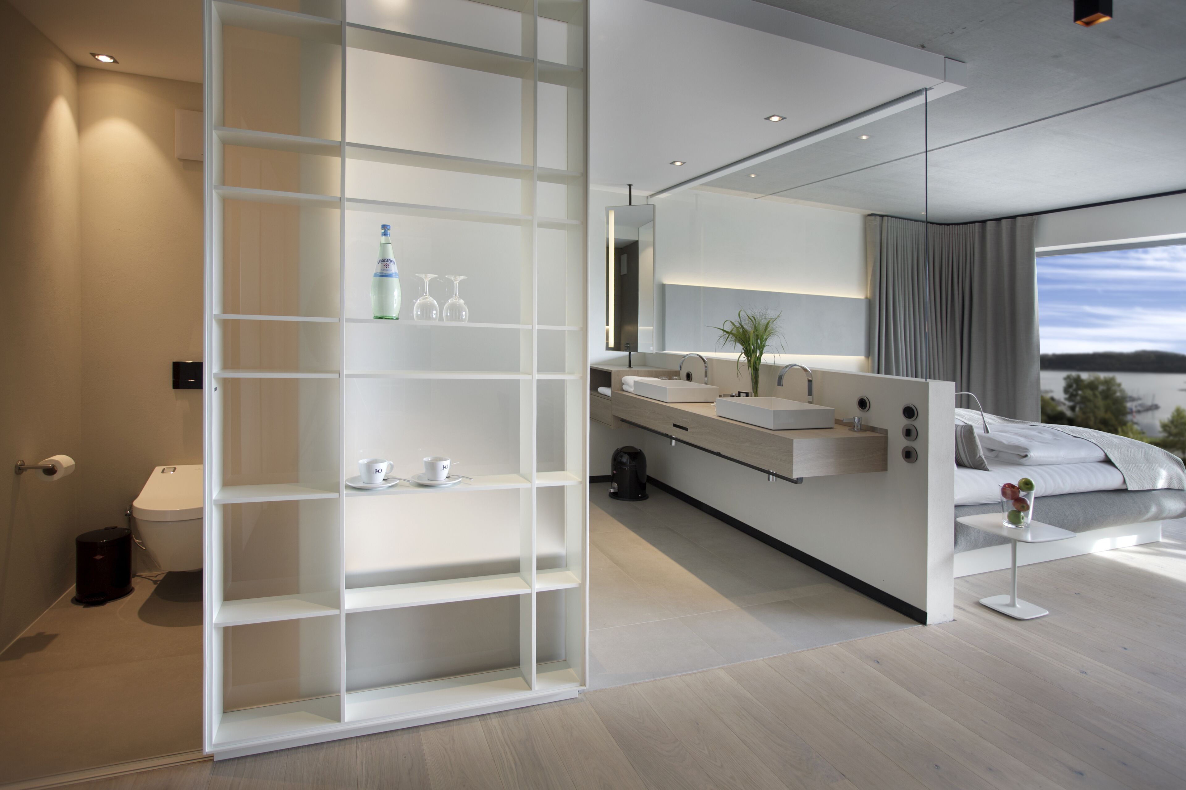 A modern bathroom with a toilet, sink, and shower area, and a bedroom with a bed and large window.