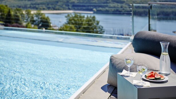 Un área junto a la piscina con una mesa para dos personas, que incluye una jarrón de vidrio, dos vasos y un plato con rebanadas de sandía.