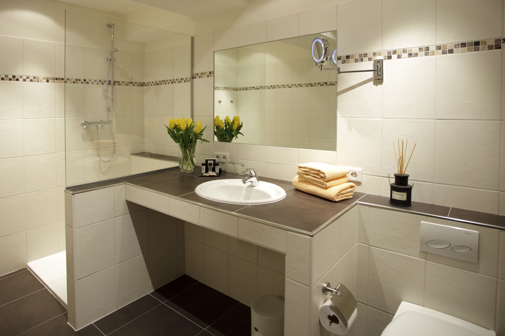 A modern bathroom with a glass-enclosed shower, a white sink, and a mirror above it. There are yellow flowers in a vase on the counter, a stack of towels, and a bottle of reed sticks.