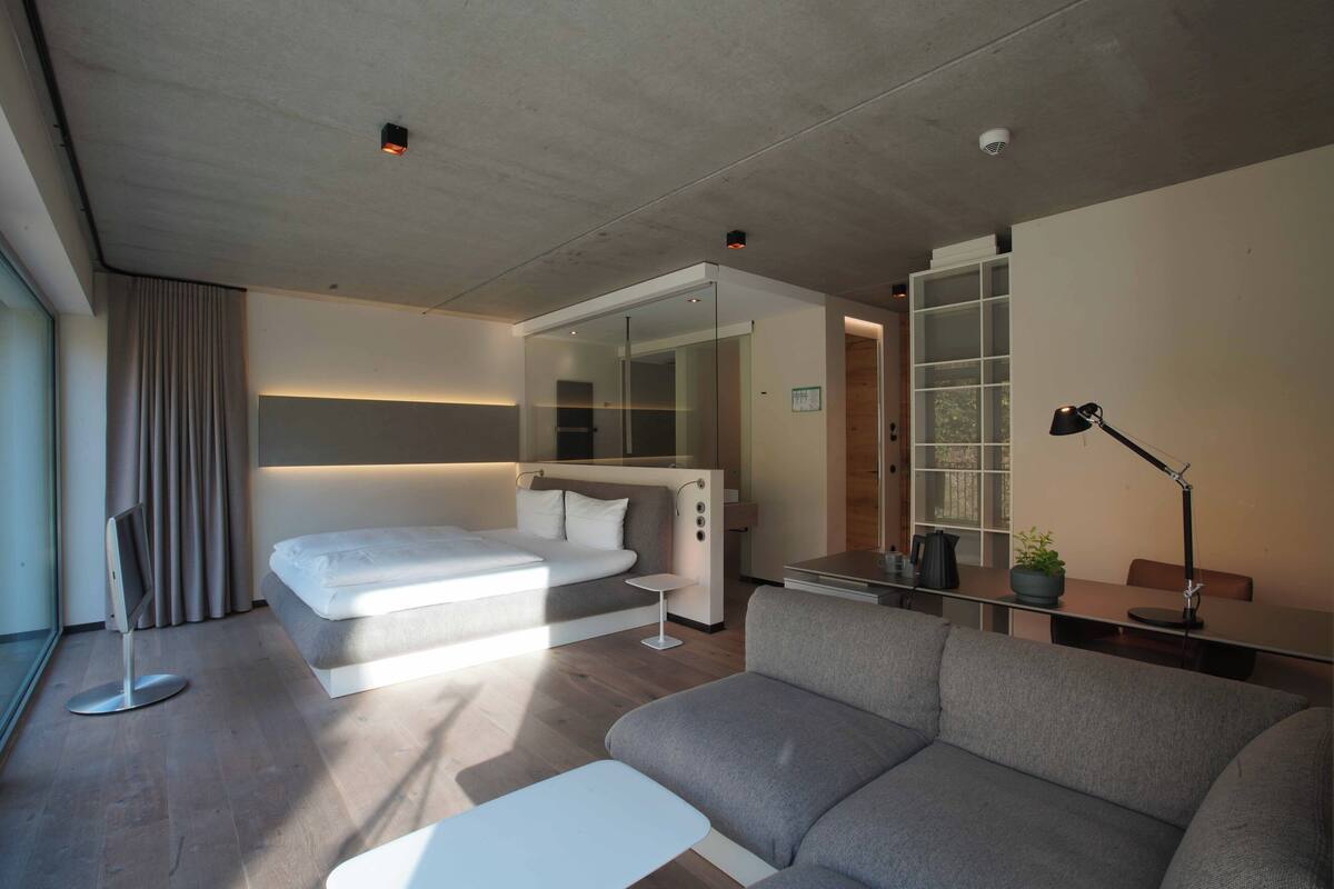 A modern hotel room with a large bed, a sofa, a desk with a lamp, and a small table.